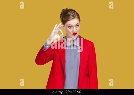 OK, va bene. La donna non dice alcun problema, mostra un gesto eccellente, ha un sorriso tenero, dà una raccomandazione, dimostra un'approvazione positiva, giacca. Studio al coperto isolato su sfondo giallo Foto Stock