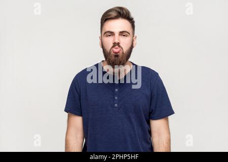 Ritratto dell'uomo con barba che indossa una T-shirt blu scuro che mostra la lingua e guarda la fotocamera con un'impertinente grimace disobbediente, che fa il volto. Ripresa da interno isolata su sfondo grigio. Foto Stock