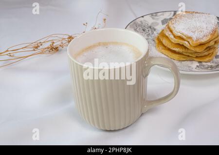 Tazza di caffè beige con vista dall'alto del latte schiumato. Sfondo bianco estetico con fiori secchi e tazza di caffè, frittelle fatte in casa d'oro, ID immagine: 2PHP8JK Foto Stock