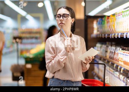Ritratto di giovane donna caucasica premurosa indossare occhiali contiene la lista dei prodotti e guardare intorno. Concetto di shopping in supermercato e consumismo Foto Stock