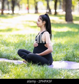 Una giovane donna incinta che conduce uno stile di vita sano e pratica yoga, medita seduta in una posizione loto su un tappetino nel parco su una calda e soleggiata mor Foto Stock