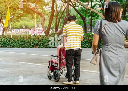 Vista posteriore dell'uomo anziano asiatico che cammina con la donna anziana disabile seduta in sedia a rotelle all'aperto indossando maschere mediche. L'uomo spinge la vecchia signora in carriola Foto Stock