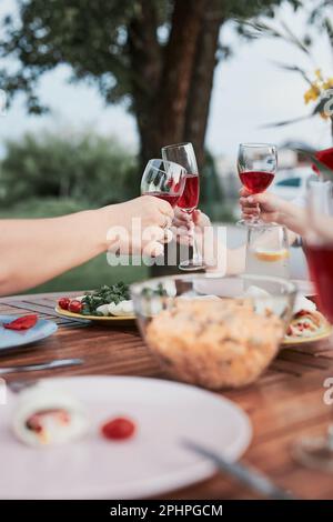 Famiglia che fa brindisi durante la cena esterna estiva in un giardino di casa. Primo piano delle mani che tengono i bicchieri di vino con vino rosso sul tavolo con i piatti Foto Stock