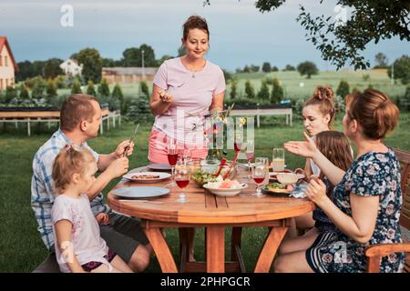 Famiglia che ha un pasto dalla griglia durante la cena estiva picnic all'aperto in un giardino di casa. Primo piano di persone sedute a un tavolo con cibo e piatti Foto Stock