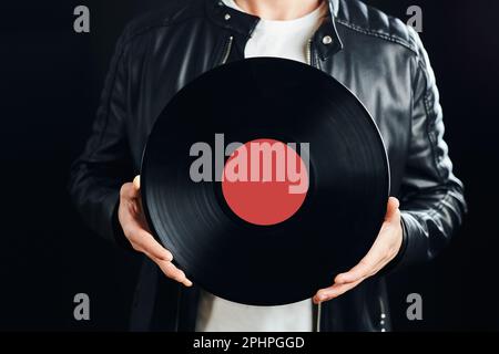 Uomo che detiene il disco in vinile. Stile musicale vintage. Maschio stile rock con giacca nera che tiene il disco nero in piedi su sfondo scuro. Musica retrò. Classe Foto Stock