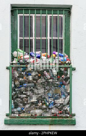 Un sacco di bottiglie di plastica e lattine scaricati attraverso barre metalliche e griglia su una finestra di un edificio commerciale. Foto Stock