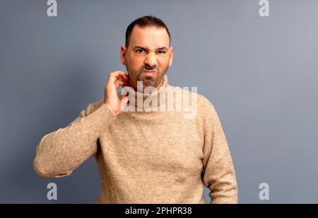 Uomo bearded di mezza età graffiandosi si è evidenziato con collare rosso su sfondo grigio studio, copia spazio. L'uomo turbato che soffre di prurito, eruzione cutanea sopra Foto Stock