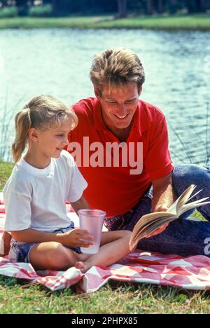 1990 PADRE BAMBINO STORICO SEDUTO AL LAGO FUORI INSEGNARE LA LETTURA INSIEME AL LAGO Foto Stock