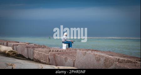 PORT ARANSAS, TX – 7 FEB 2023: Pescatore anziano seduto pazientemente su un secchio di plastica mentre pesca al South Jetty a Port Aransas, Texas. Foto Stock