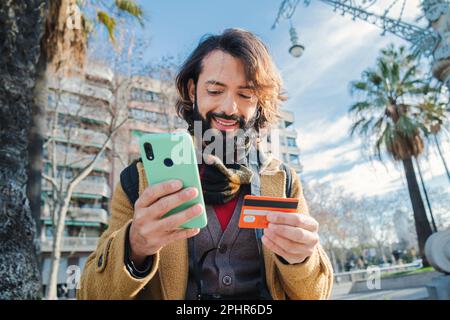 giovane uomo che paga un acquisto con una carta di credito e che tiene un dispositivo smartphone all'aperto. Felice uomo bearded utilizzando un cellulare app per l'acquisto di una consegna e guardare la finanza bancario. Foto di alta qualità Foto Stock