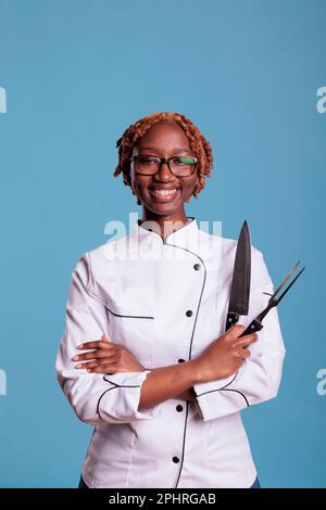 Cucina afro-americana femminile, capelli ricci posa con coltello da cucina professionale, affilatrice. Chef sorridente che tiene utensili da cucina. Ritratto di giovane lavoratrice di ristorazione in uniforme. Foto Stock
