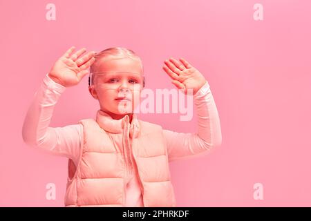 Ragazza con abiti futuristici e occhiali vr sta sperimentando il cyberspazio con luce al neon rosa su sfondo rosa. Foto Stock