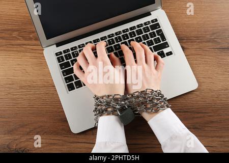 Donna con mani incatenate che digita sul laptop al tavolo di legno, vista dall'alto. Dipendenza da Internet Foto Stock