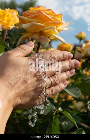 Mani piegate da donna con un rosario illuminato dal sole sullo sfondo di rose gialle e cielo blu, primo piano. Foto Stock