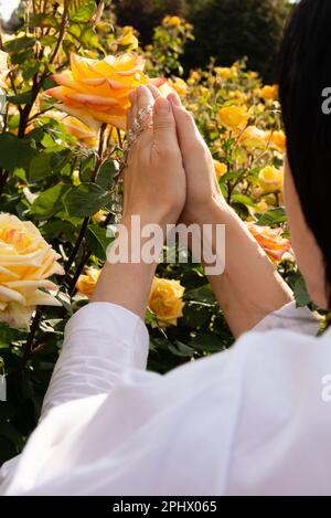 Vista a spalla di una donna in preghiera con un rosario in mano sullo sfondo di rose gialle in una giornata di sole. Foto Stock