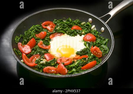 Uovo fritto con spinaci e pomodori in una padella su un piano cottura nero, la preparazione di un pasto sano per la colazione o il pranzo, spazio copia, focus selezionato Foto Stock