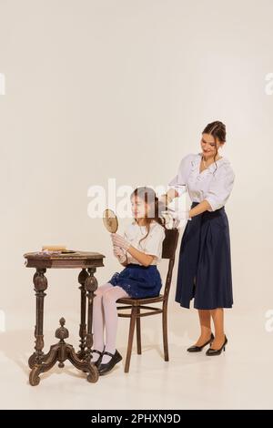 Ritratto di giovane donna che indossa abiti antiquati intrecciando i capelli della figlia e sorridendo su sfondo chiaro Foto Stock