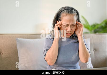 Stressata e malata donna asiatica si siede sul divano nel suo salotto, toccando i suoi templi e soffrendo di mal di testa o emicrania. Problemi di salute Foto Stock