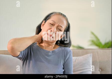 Donna asiatica anziana infelice siede sul divano nel suo salotto che soffre di dolore al collo e sfrega il collo. Foto Stock