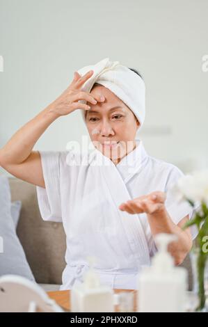 Ritratto di bella donna asiatica in accappatoio applicando siero viso sul viso dopo la doccia. Foto Stock