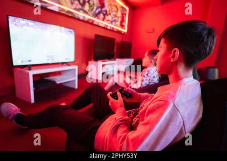 Due giocatori ragazzi giocano a calcio gamepad console di videogiochi nella sala giochi rossa. Foto Stock