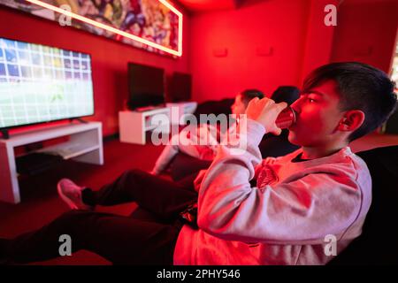 Due giocatori ragazzi giocano a calcio gamepad console di videogiochi nella sala giochi rossa. Lattina di soda da bere. Foto Stock