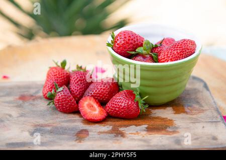 Elegante ciotola verde con fragole sdraiate su una tavola bagnata all'aperto durante il sole. Foto Stock