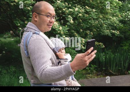 Un nuovo padre sorridente scattando una foto con il suo telefono delle cellule mentre trasporta il suo bambino maschio con un portatore del bambino nella natura al parco nella stagione primaverile. Brit Foto Stock