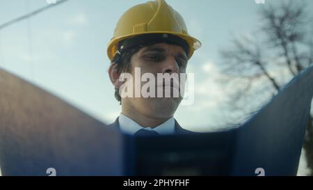 Ingegnere o appaltatore di costruzione senza barba in un casco e vestito che esamina Blueprints in una cartella blu e che ottiene frustrato sopra un dettaglio difettoso Foto Stock