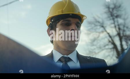 Un giovane, ingegnere o appaltatore di costruzione senza orecchio in un casco e vestito che esamina i Blueprints in una cartella blu e che ottiene frustrato sopra un difettoso Foto Stock