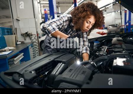 La giovane donna in una camicia a quadri ispeziona attentamente l'auto dal cappuccio Foto Stock