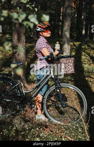 Donna attiva che trascorre il tempo libero delle vacanze estive in un viaggio in bicicletta in una foresta. Donna che indossa il casco e i guanti per bicicletta con cesto e dr Foto Stock