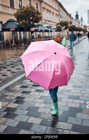 Bambina che si nasconde dietro un grande ombrello rosa che cammina in un centro in un giorno d'autunno piovoso e cupo Foto Stock