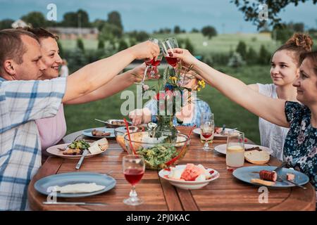 La famiglia fa brindisi durante la cena estiva picnic all'aperto in un giardino di casa. Primo piano di persone che tengono bicchieri da vino con vino rosso sul tavolo con cibo A. Foto Stock