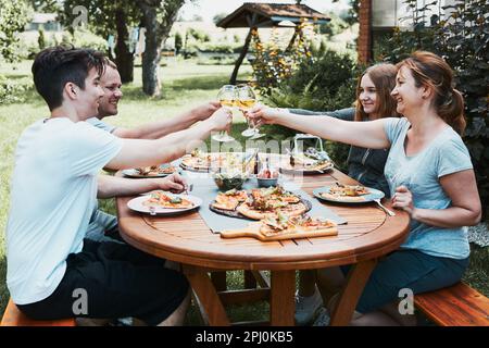 Gli amici fanno brindisi durante il picnic estivo cena all'aperto in un giardino di casa. Primo piano di persone che tengono bicchieri da vino con vino bianco sul tavolo con piz Foto Stock