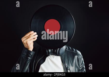 Uomo che si nasconde dietro la testa di copertura del disco in vinile retro. Stile rock. Stile musicale vintage. Uomo che indossa una giacca nera che tiene il disco in vinile in piedi sul nero Foto Stock