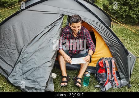 Uomo che guarda la mappa mentre pianifica il prossimo viaggio. Uomo che si rilassa in tenda in campeggio durante le vacanze estive. Trascorrere attivamente vacanze all'aperto vicino a nat Foto Stock