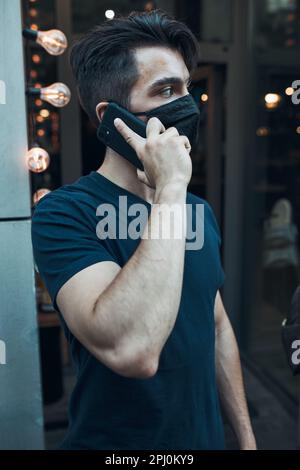 Giovane uomo in piedi di fronte al ristorante che parla al telefono utilizzando lo smartphone indossare la maschera per evitare l'infezione da virus e per prevenire la diffusione o Foto Stock