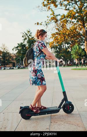 Giovane donna in sella a uno scooter elettrico nel centro della città. Donna che indossa la maschera per evitare l'infezione da virus e per prevenire la diffusione della malattia i Foto Stock