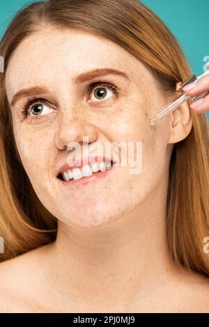 Ritratto di donna sorridente frettolata applicando siero cosmetico isolato sul turchese, immagine stock Foto Stock