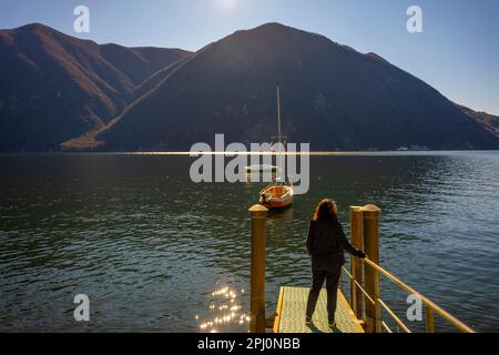 Donna in piedi su un molo con barca a vela sul Lago di Lugano e sulla catena montuosa in una giornata di sole con cielo limpido a Lugano, Ticino, Svizzera. Foto Stock