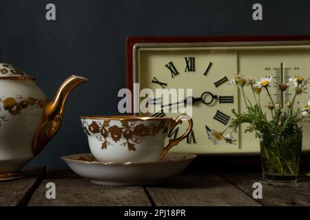 una tazza di latte e un vaso con margherite vicino all'orologio su un tavolo di legno Foto Stock