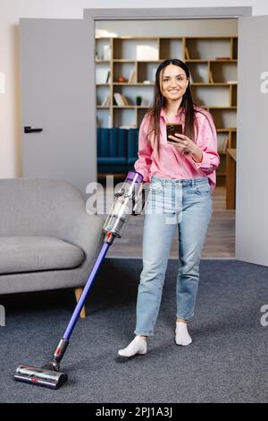 Felice giovane caucasica bella donna vacuuming pavimento e SMS, navigare su Internet su smartphone a casa in soggiorno. Abbastanza femmina utilizzando Foto Stock