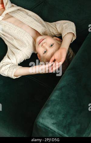 Ritratto di un ragazzo in un accappatoio beige, che giace su un divano verde e guarda la macchina fotografica. Foto Stock
