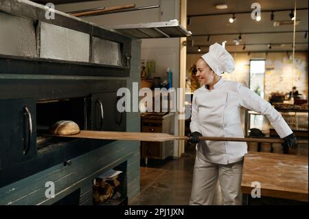 Giovane lavoratore di panetteria usando pala di legno per prendere il pane dal forno di stufa Foto Stock