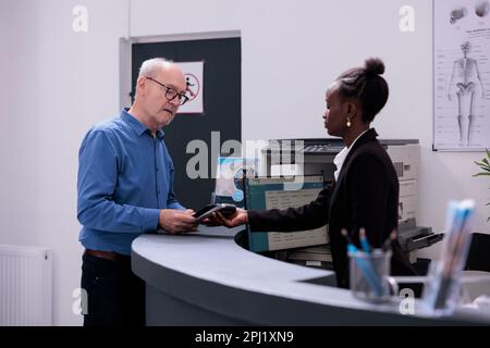 Paziente anziano in piedi presso il banco di ricevimento dell'ospedale che paga un esame medico con lo smartphone utilizzando senza contatto, parlando con il receptionist nella lobby. Vecchio che effettua il pagamento dopo aver consultato Foto Stock