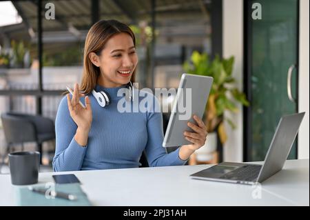 Donna asiatica sorridente e felice, freelancer in abiti casual, che parla con il suo cliente in una videochiamata tramite tablet in uno spazio di co-working. Foto Stock