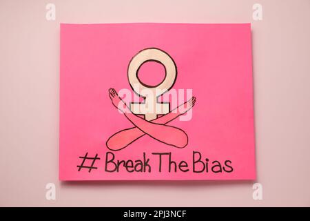 Carta con hashtag BreakTheBias, simbolo del genere femminile e disegno di braccia incrociate su sfondo rosa, vista dall'alto Foto Stock