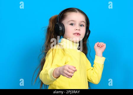 Felice bambina con cuffie wireless su sfondo blu che fa la danza si muove verso la musica in cuffie, bambino con capelli lunghi. Foto Stock