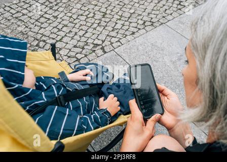 La mamma usa un messaggero sul suo telefono cellulare vicino al passeggino con il figlio che dorme. Foto Stock